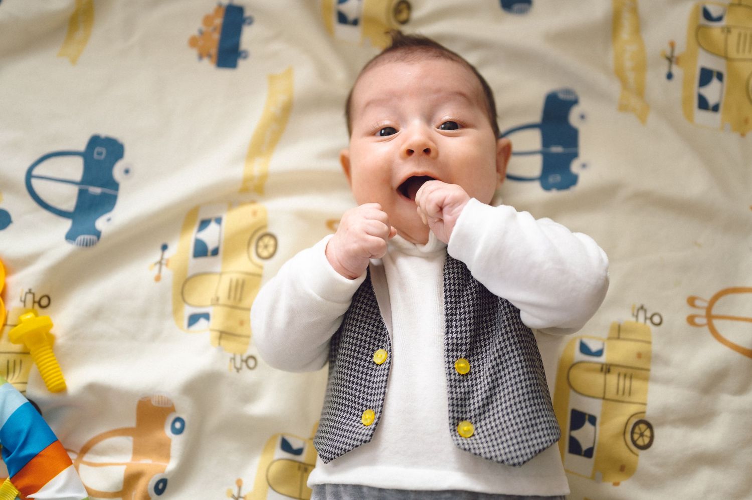 Baby in white and grey outfit lying on colorful patterned blanket with vehicle designs.