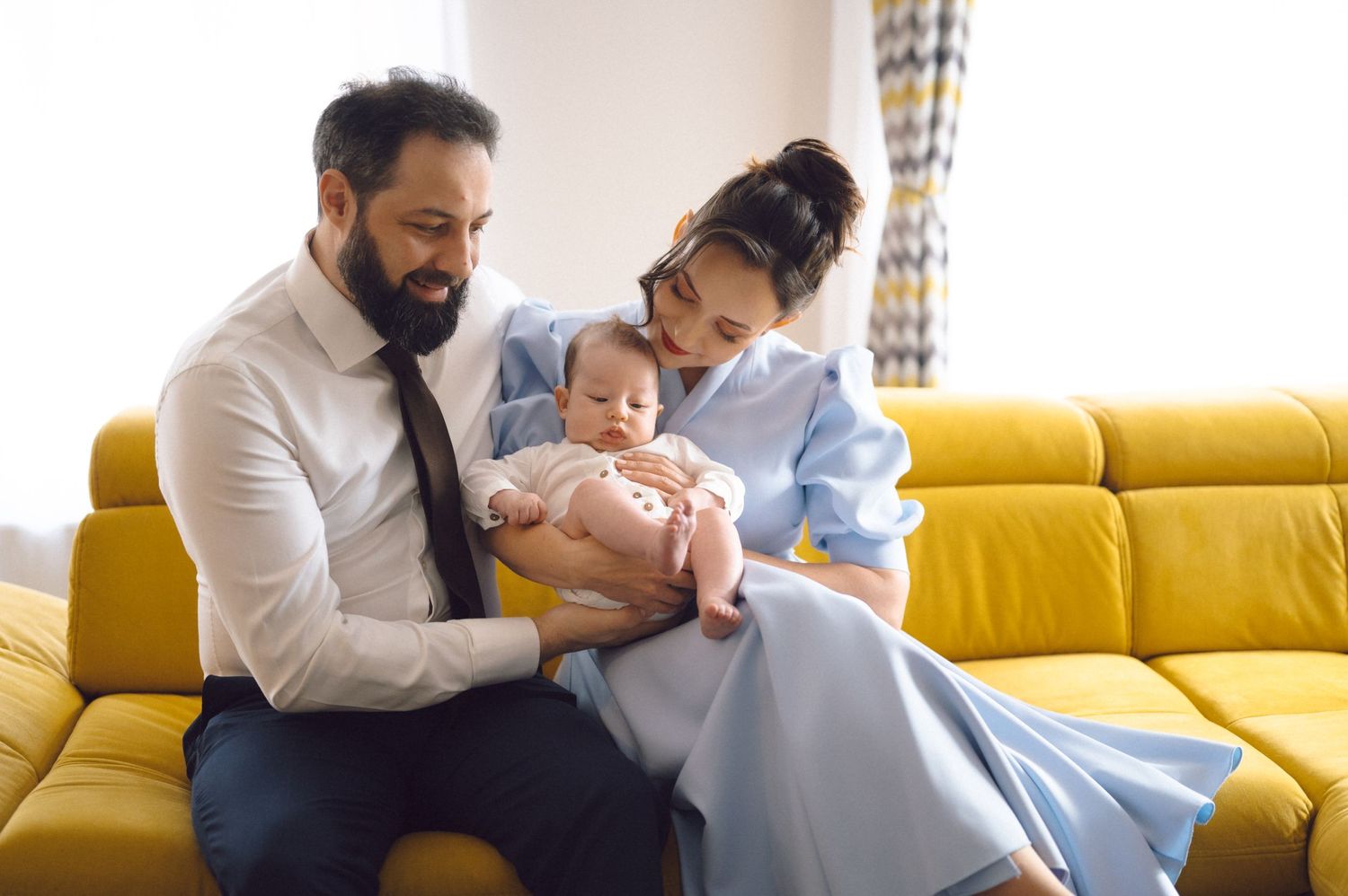 A couple sitting on a yellow couch while cradling their baby.