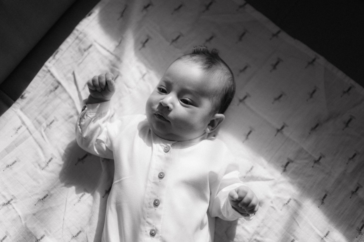A baby in a white onesie lying on patterned fabric, captured in black and white.