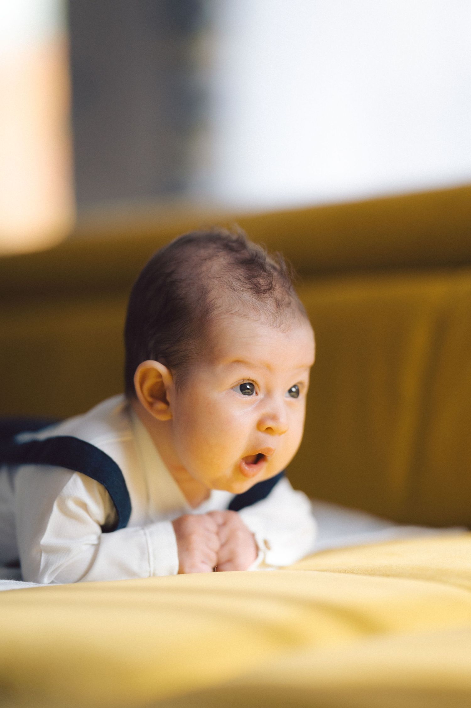 A baby in white clothing looking up while laying on a yellow surface.