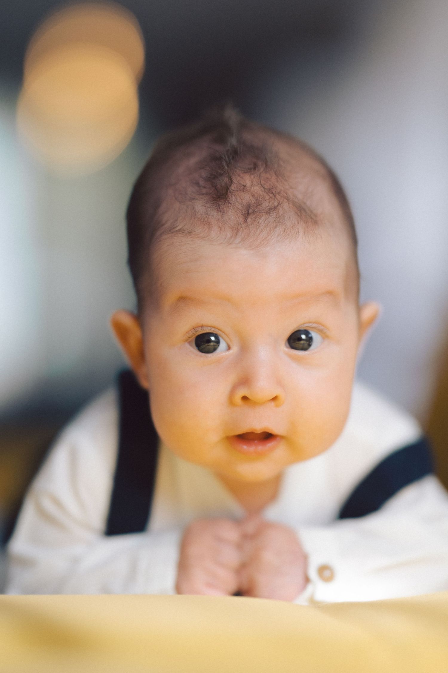 A baby in white clothing and dark suspenders looking at camera with wide eyes.