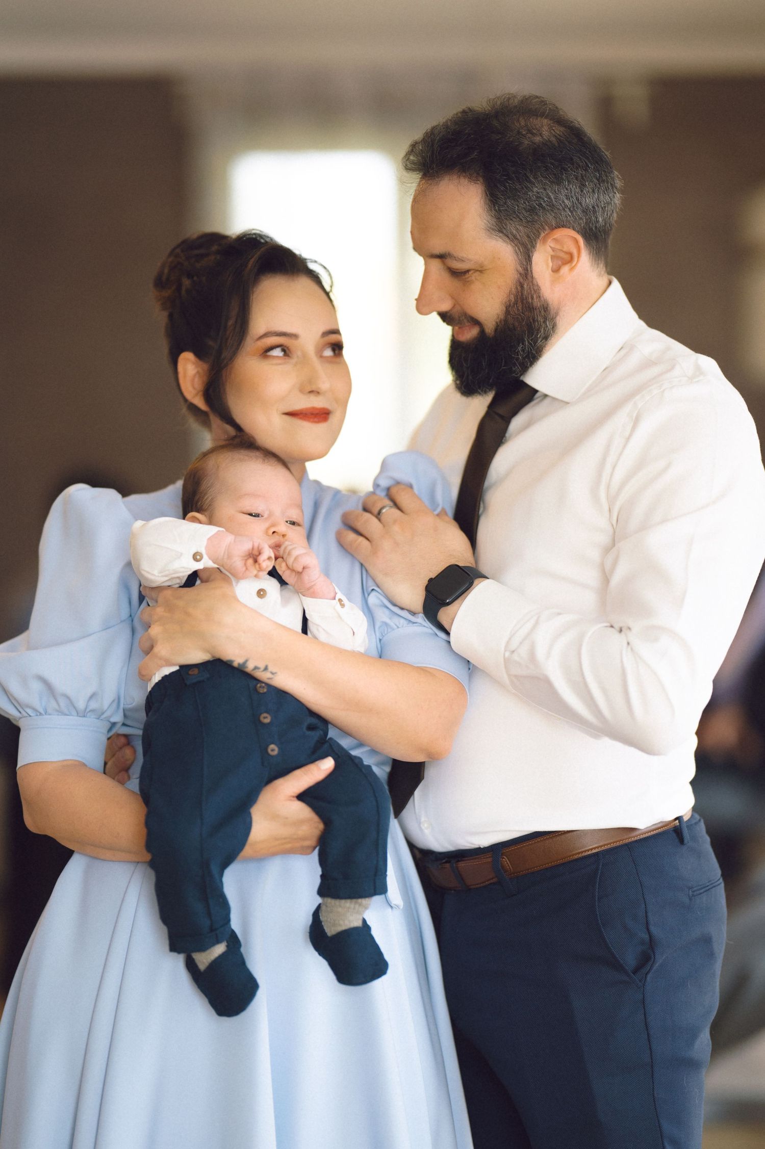 A couple in formal attire holds their infant child while sharing an intimate family moment.