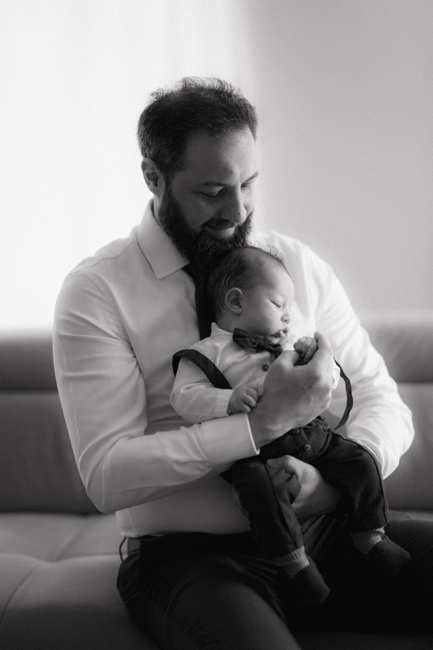 A bearded man in a white dress shirt cradles a sleeping baby on a gray couch.