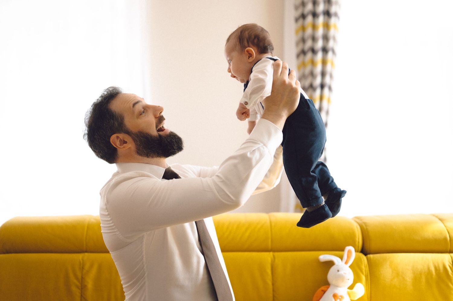 A father in a white shirt playfully lifts his baby in the air near a yellow couch.