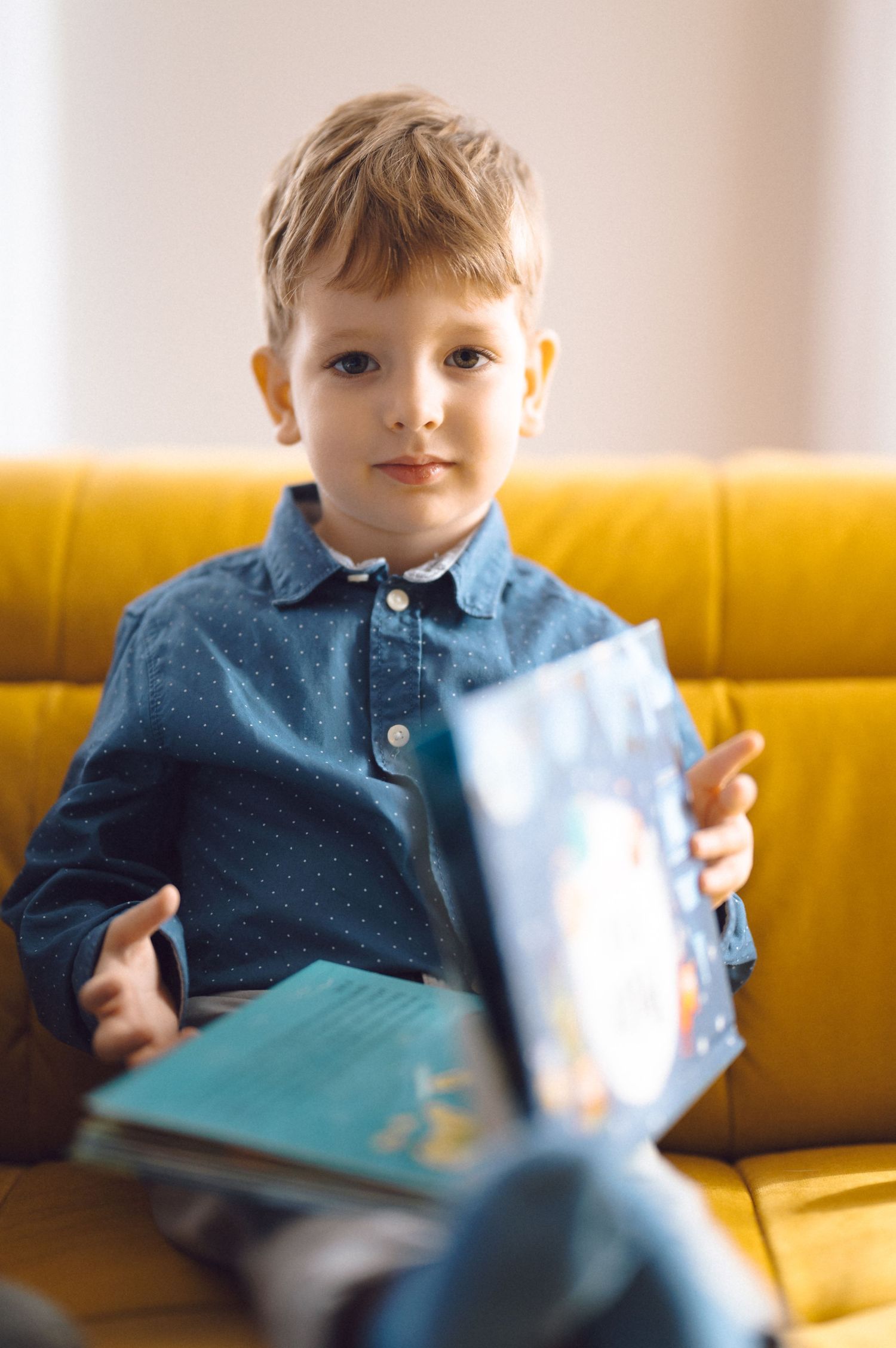 Young boy in blue polo shirt sits on yellow couch reading a book.