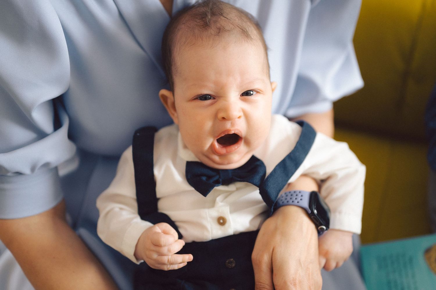 Smiling baby wearing formal outfit with bow tie sits on yellow couch.