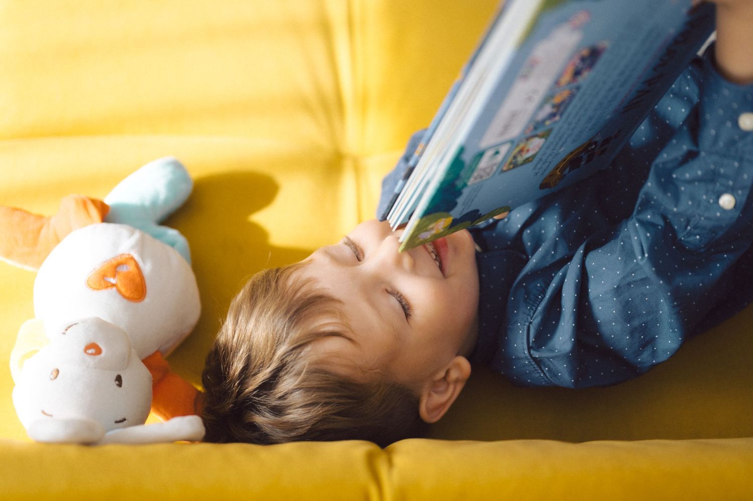 Child lying on yellow couch reading book with stuffed toy nearby.