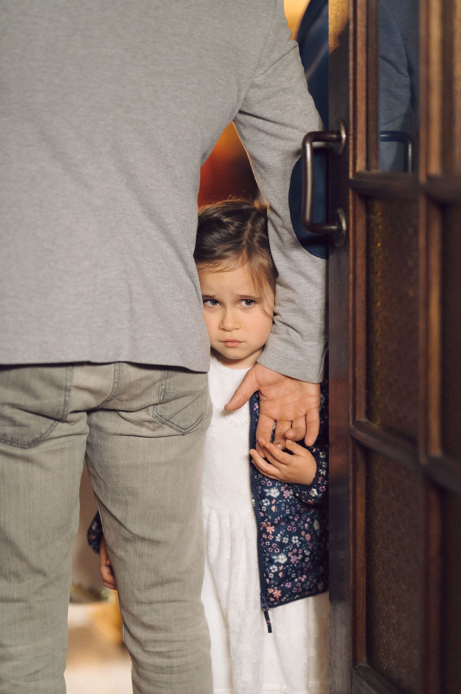 Child holding an adult's hand while partially hidden behind their legs in a timid pose.