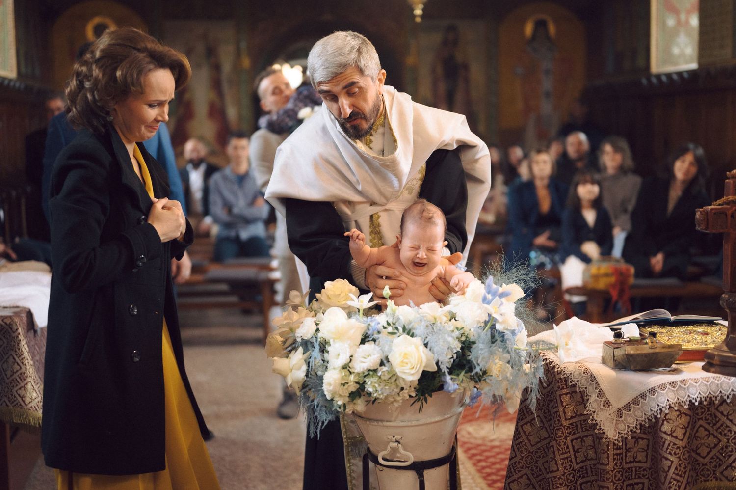 Orthodox baptism ceremony with white floral arrangements in a traditional church setting.