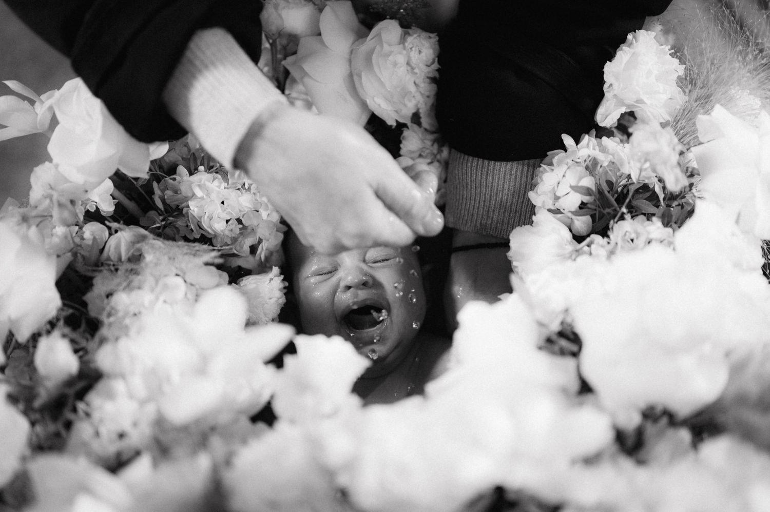 Black and white image of a baby surrounded by white flowers during a baptism.