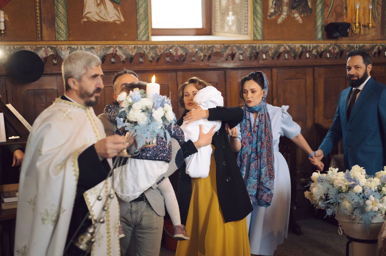 Religious ceremony with priest and family members surrounded by white floral arrangements.