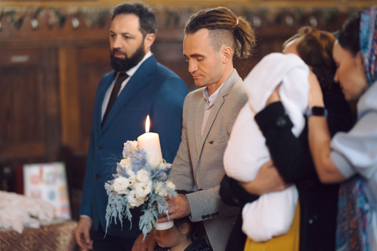 Two people stand in a church ceremony with a decorated white candle and floral arrangements.