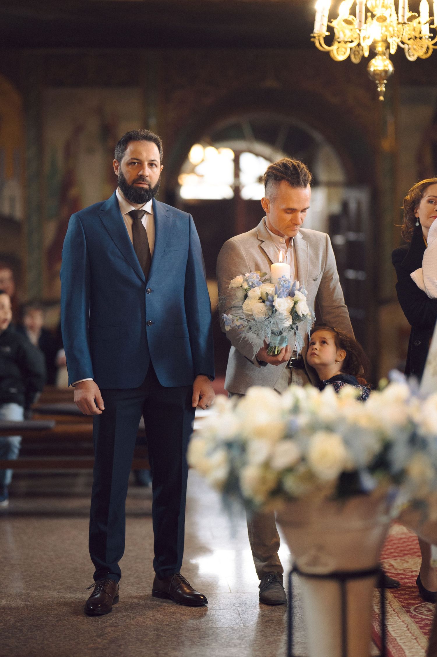 Two adults and a child stand in an ornate church interior with floral decorations and a chandelier.