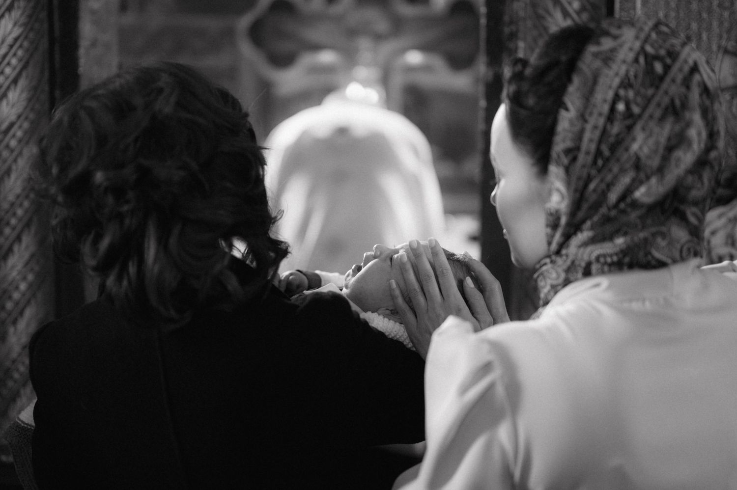 A black and white photograph shows a baptism ceremony with a priest and family members.
