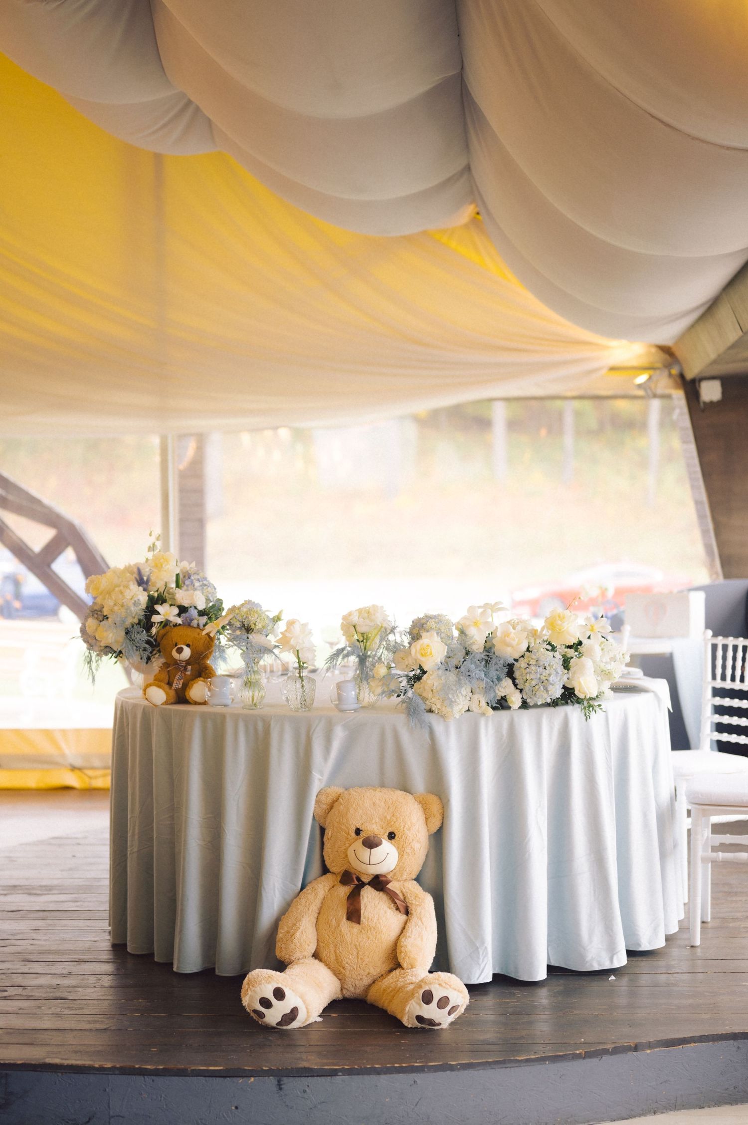 Wedding reception table with flowers and teddy bear under draped white tent ceiling.