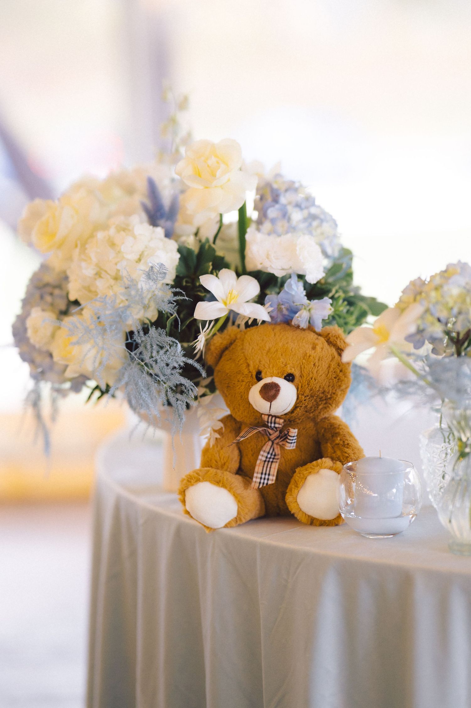 Teddy bear nestled among yellow and blue floral arrangements on white tablecloth.