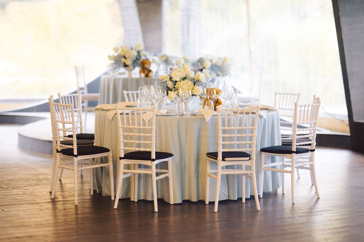 Wedding reception hall with white chiavari chairs and floral centerpieces on round tables.