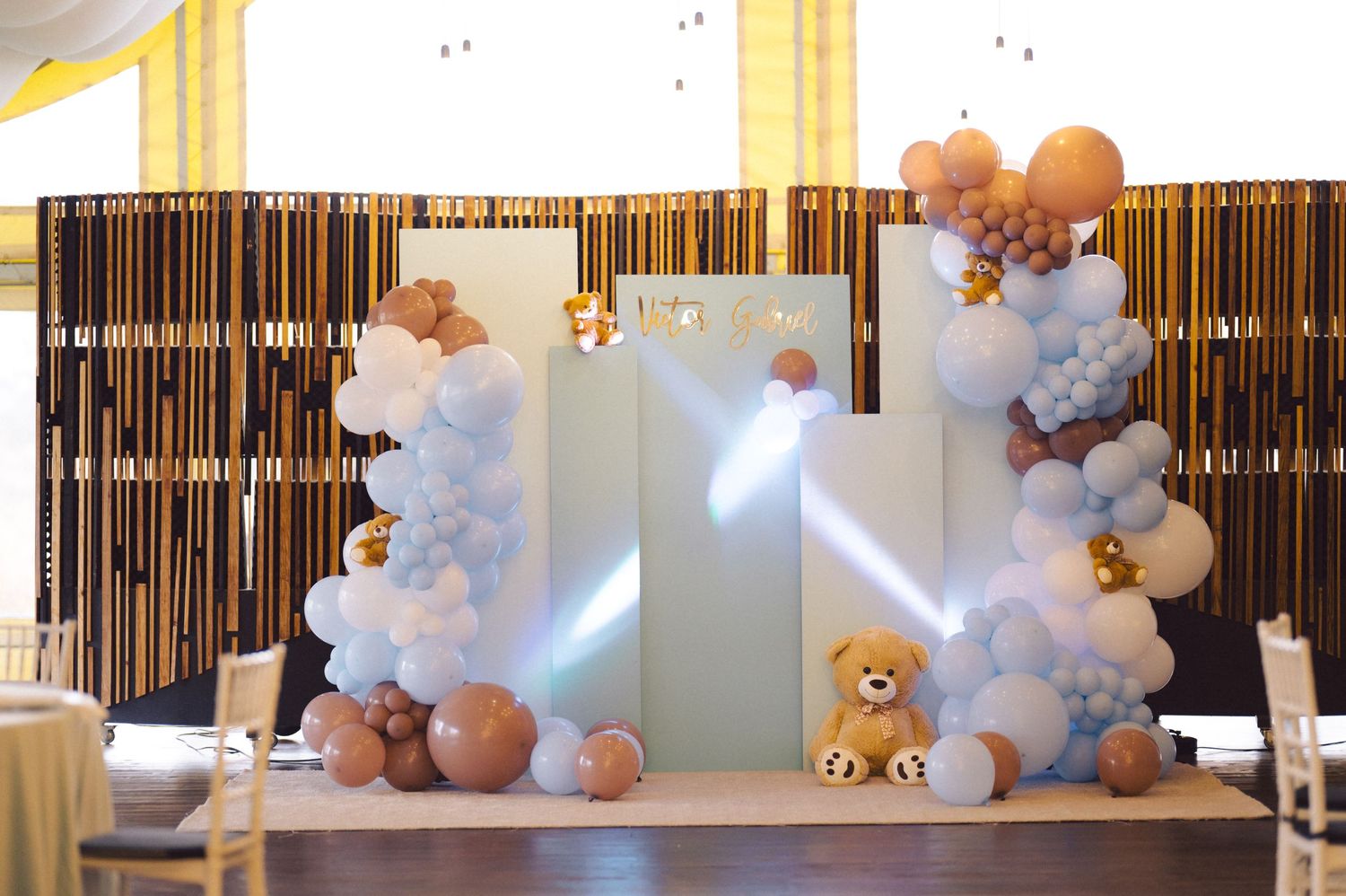Teddy bear themed balloon arch with blue and brown decorations against bamboo backdrop.