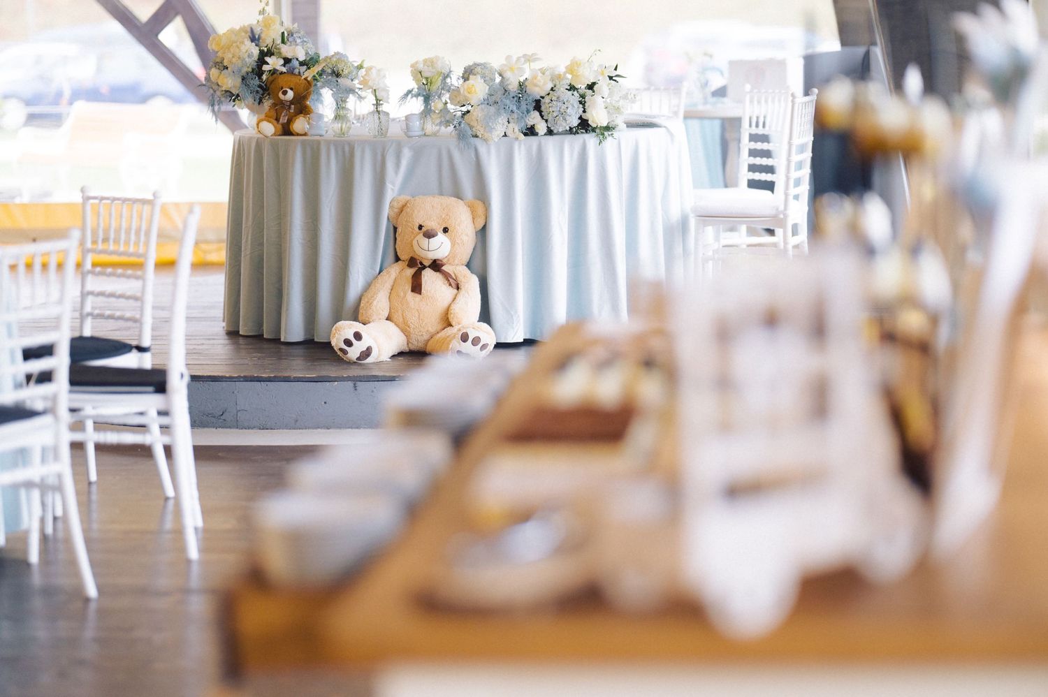 Teddy bear sits near an elegant white tablescape with floral arrangements at a reception venue.