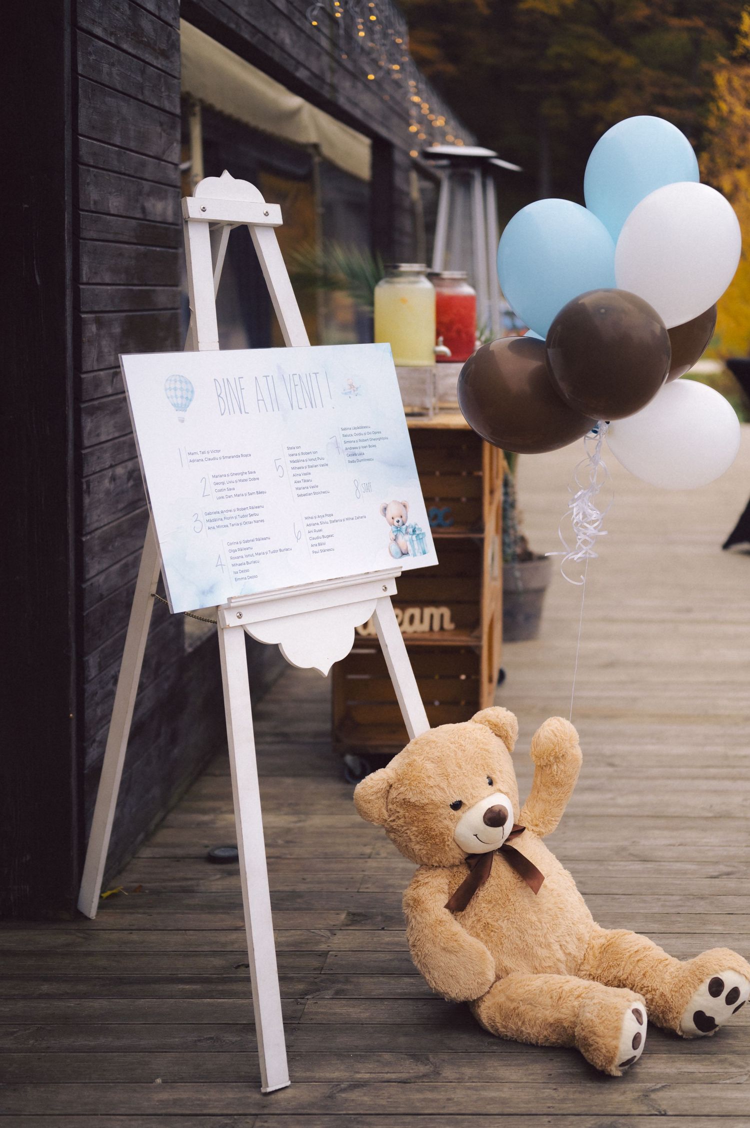 Teddy bear and balloons next to white easel with sign on wooden deck.