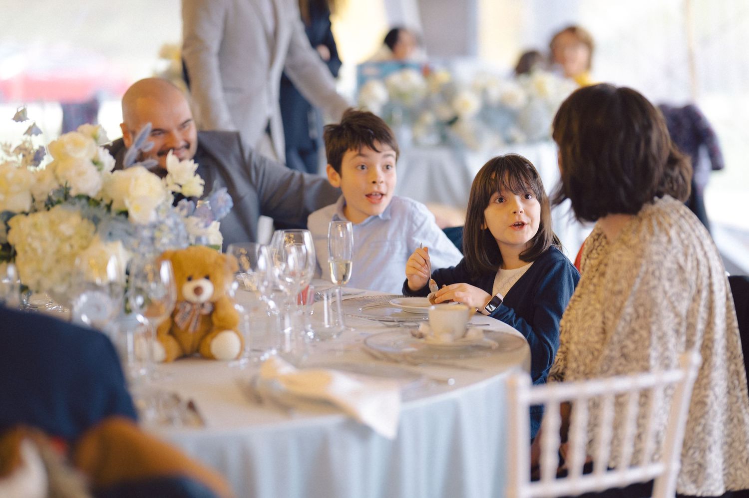 Event guests seated at round tables with teddy bear centerpieces and white linens.