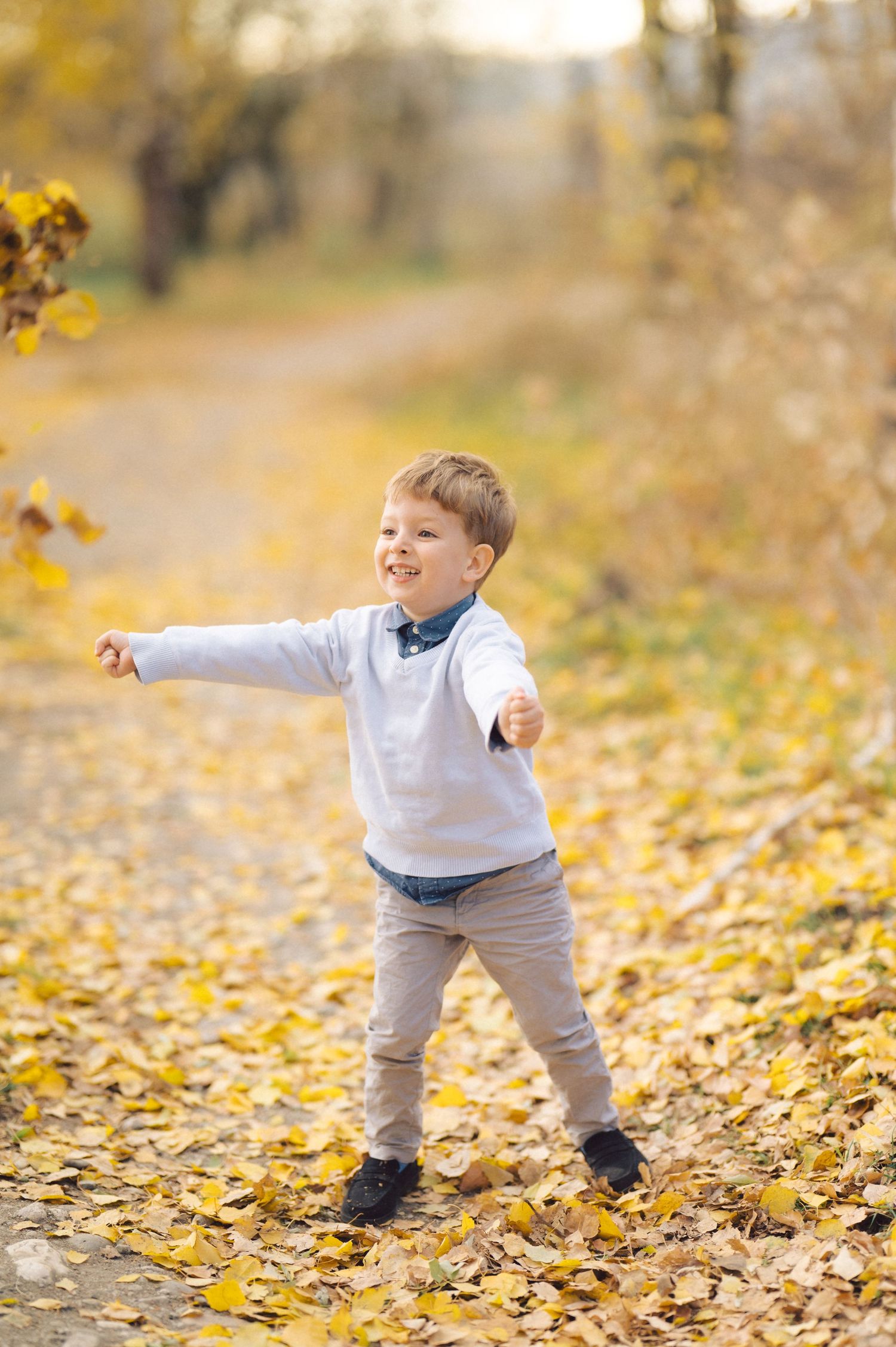 Young child plays in fallen autumn leaves wearing a white sweater and grey pants.