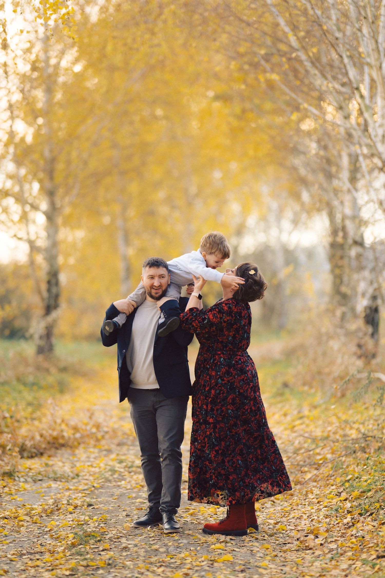 A family enjoys a playful moment on a tree-lined path covered in yellow autumn leaves.