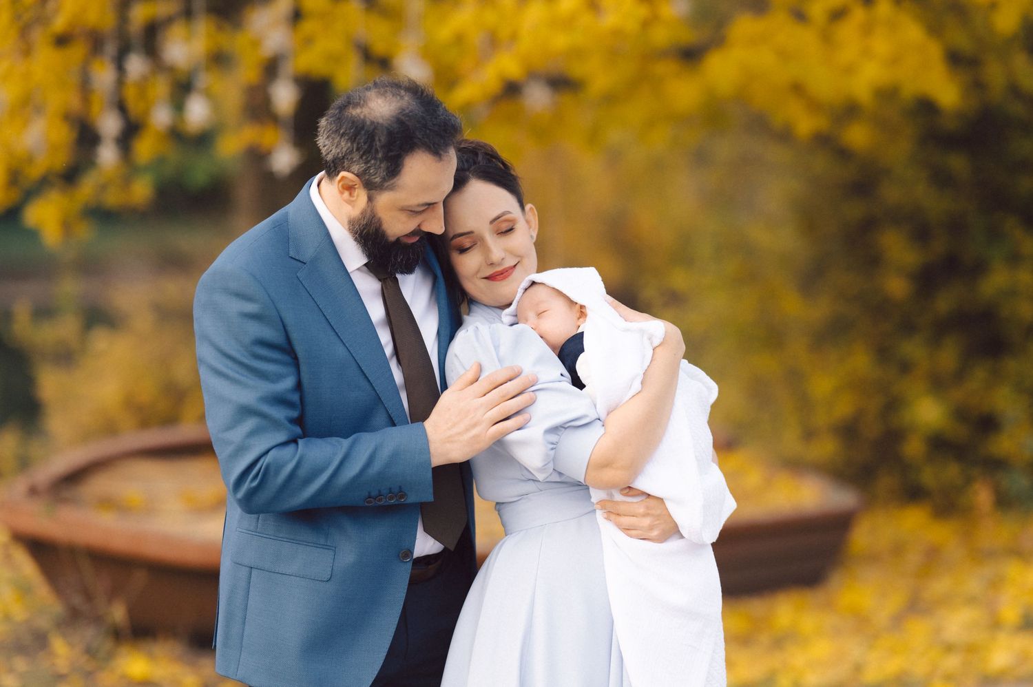 A couple embraces tenderly while holding a newborn baby against a backdrop of golden autumn trees.