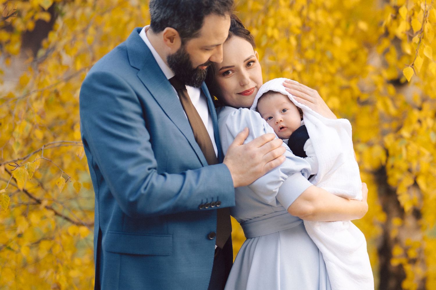 A family shares a tender moment with their newborn baby against yellow autumn leaves.