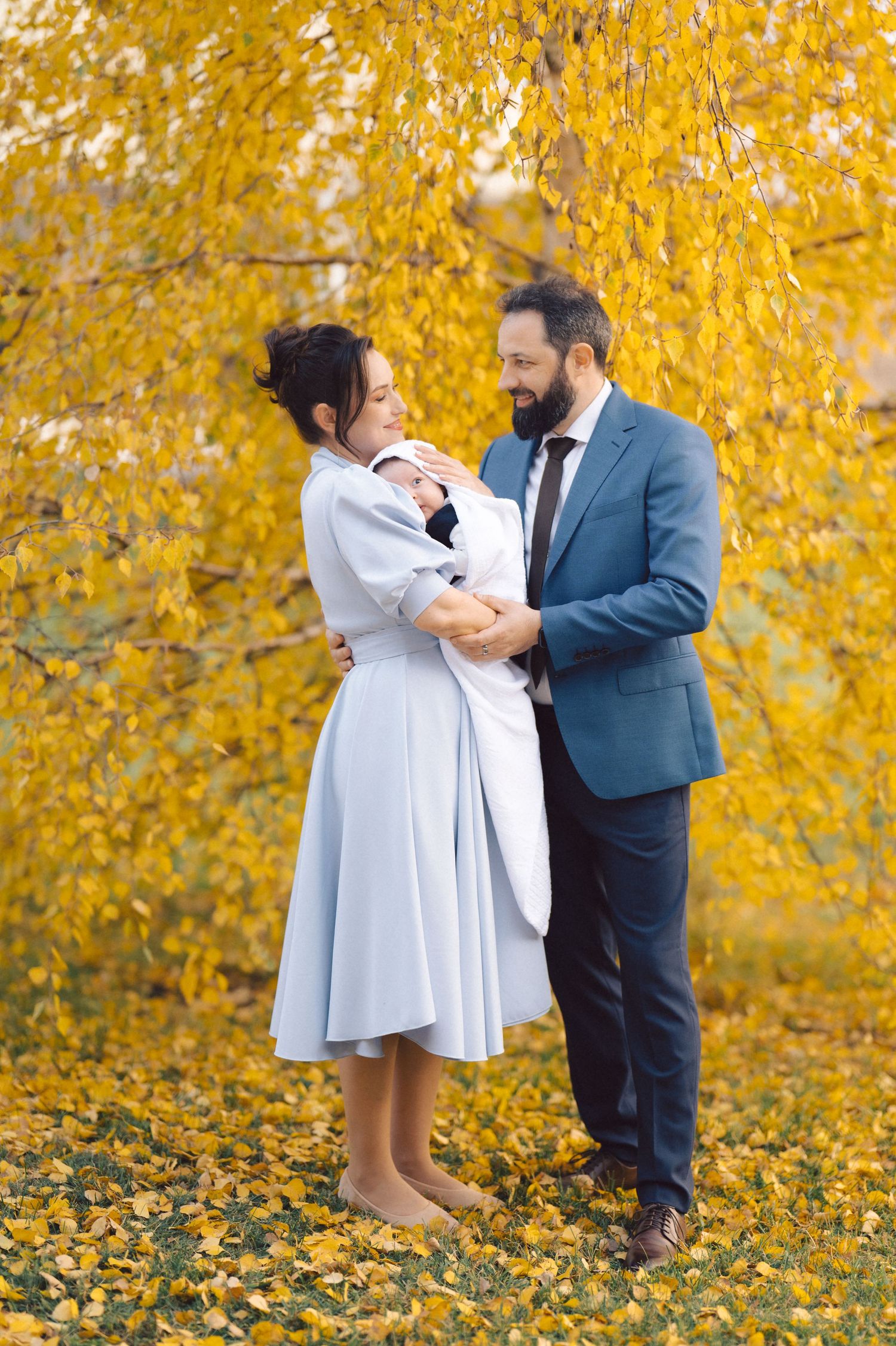 A couple dances together while holding their baby beneath golden autumn leaves.
