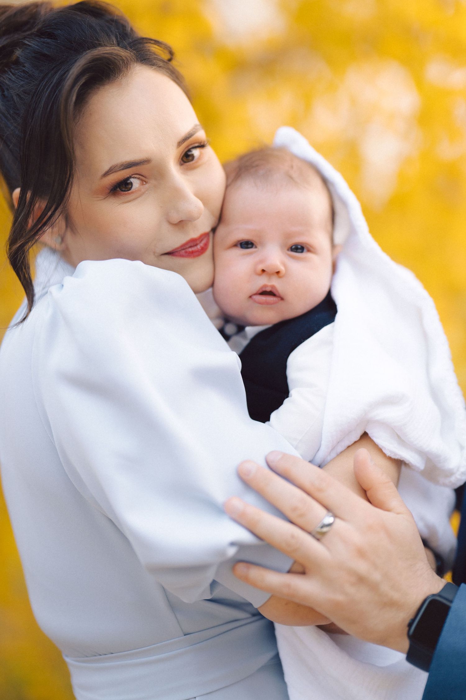 Baby wearing white outfit is held lovingly against autumn yellow background.