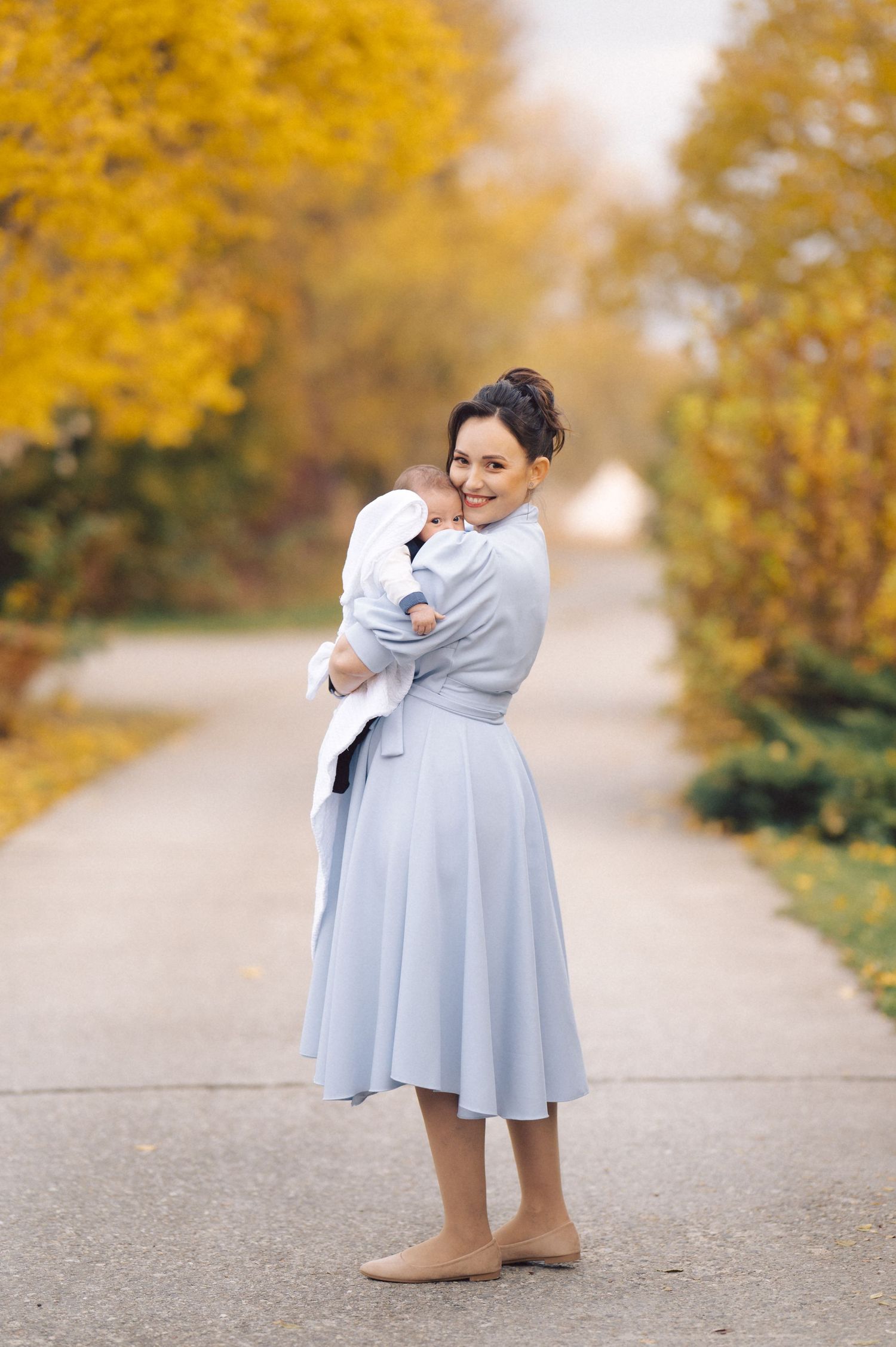 Woman in light blue dress holds baby on autumn path with golden trees.