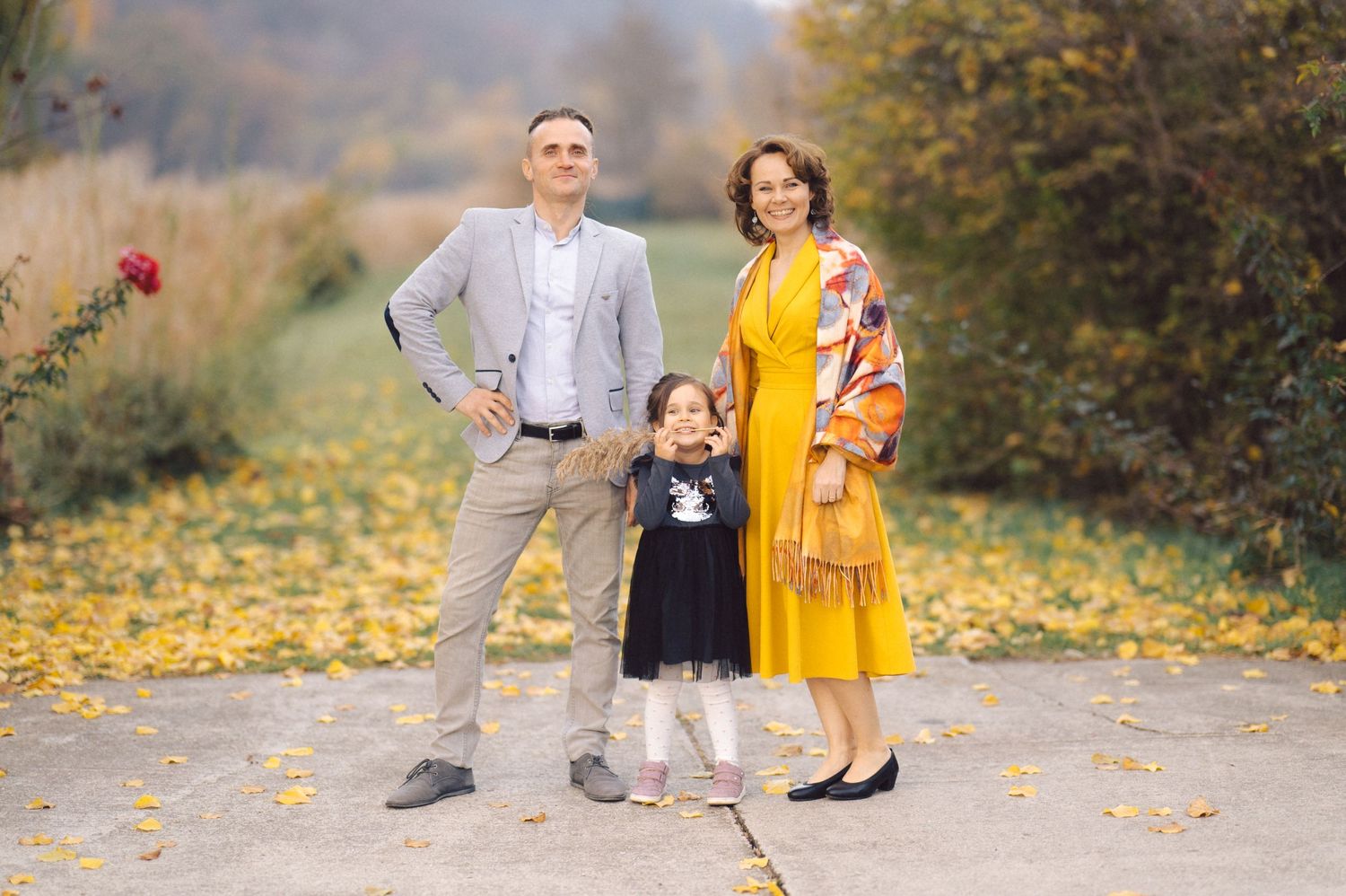Family of three stands together on an autumn path covered in yellow leaves wearing complementary yellow and gray outfits.
