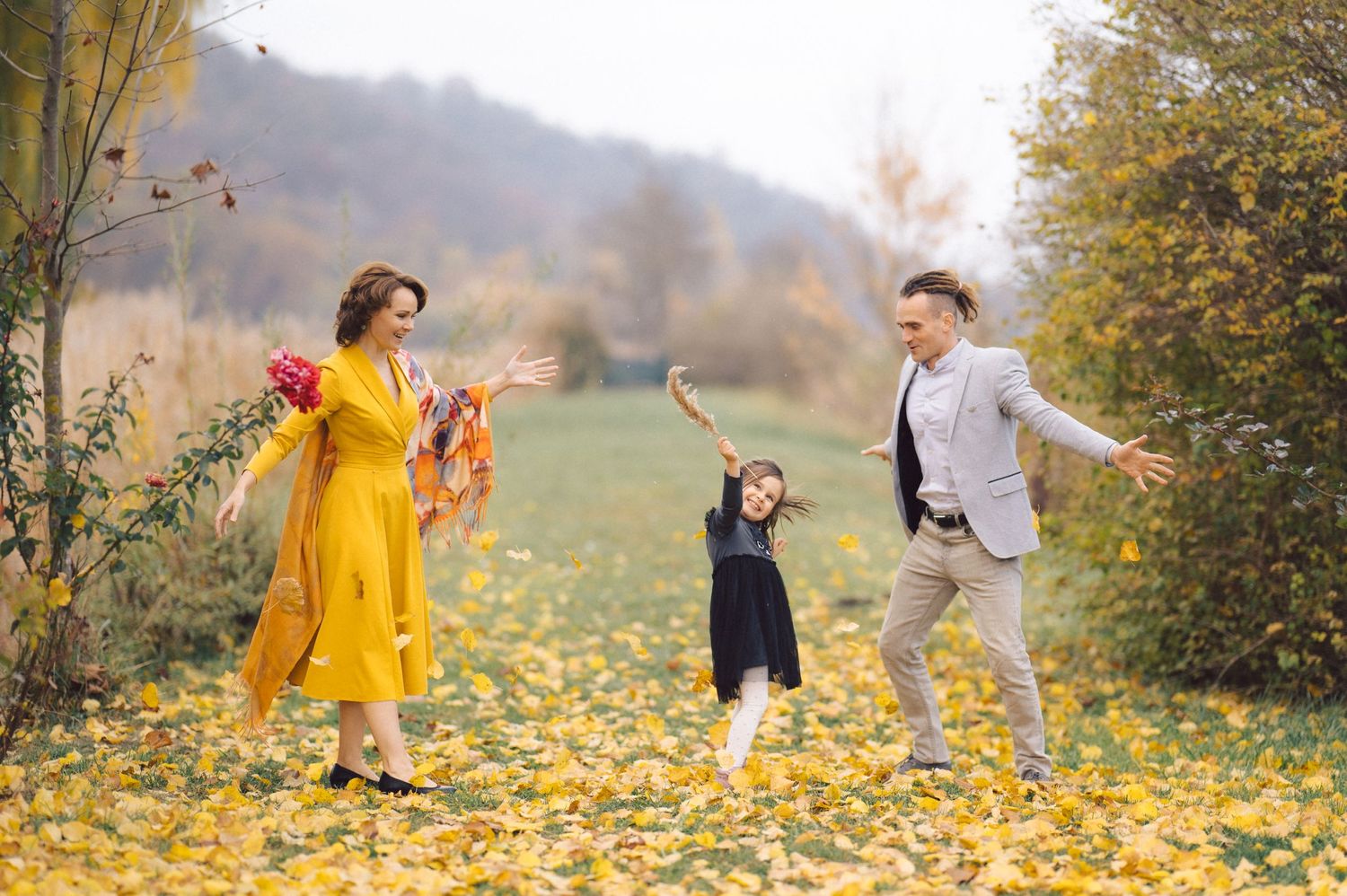 Family plays together in a field of fallen autumn leaves with rolling hills in background.