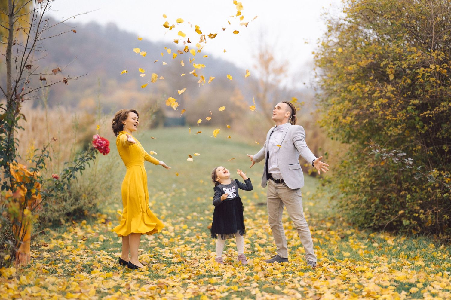 Family enjoys playful moment tossing fallen leaves in autumn scenery with mountains behind.