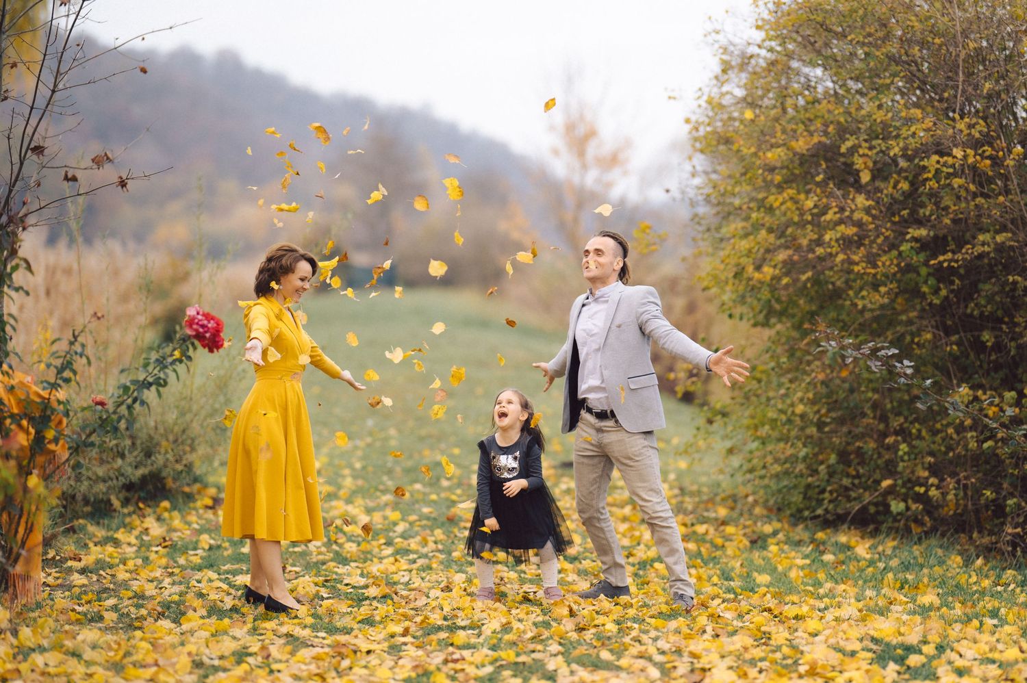 Family playing with fallen leaves on ground during autumn photoshoot in nature.