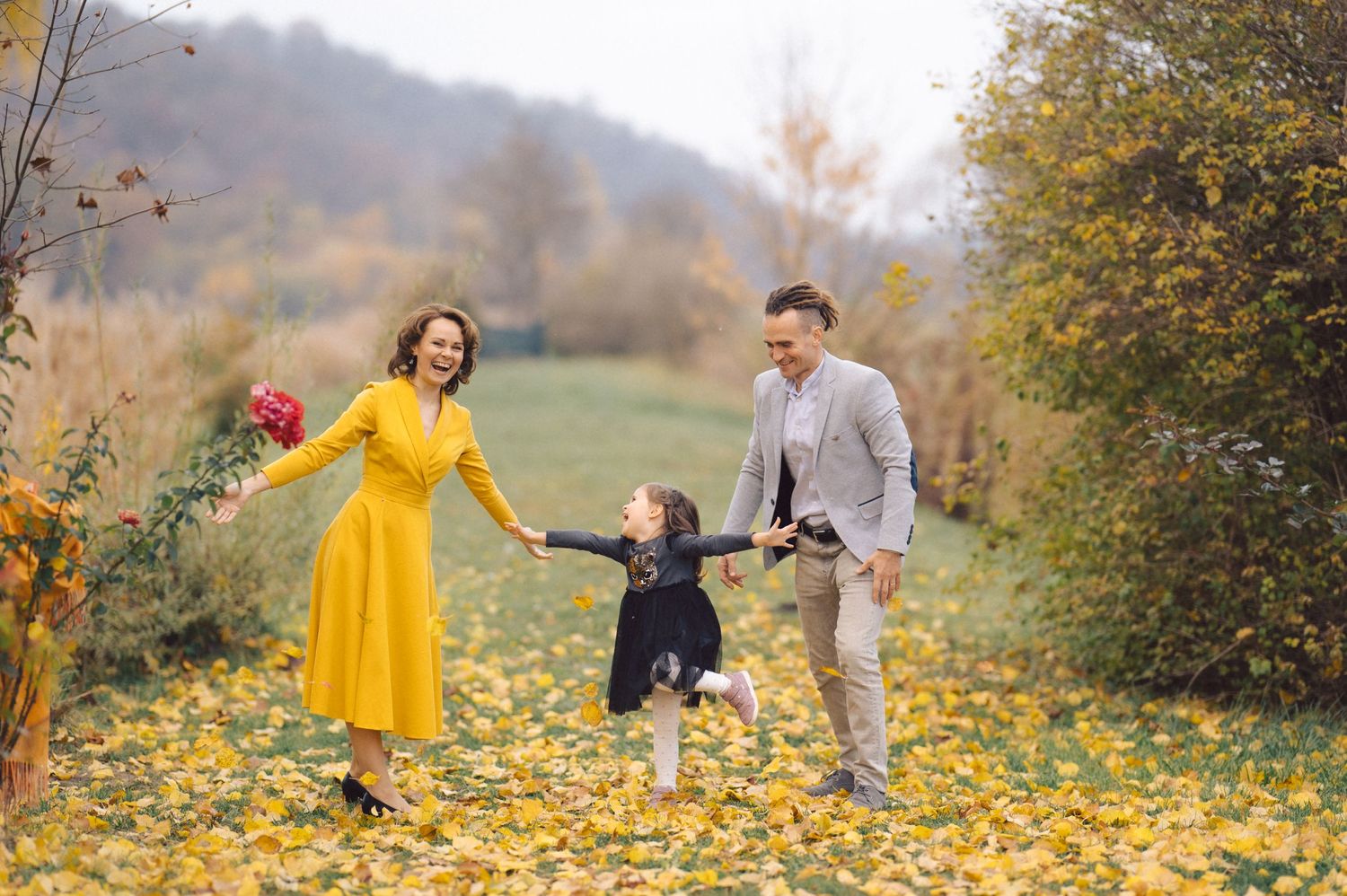 Family holds hands while walking through yellow fallen leaves in autumn landscape.