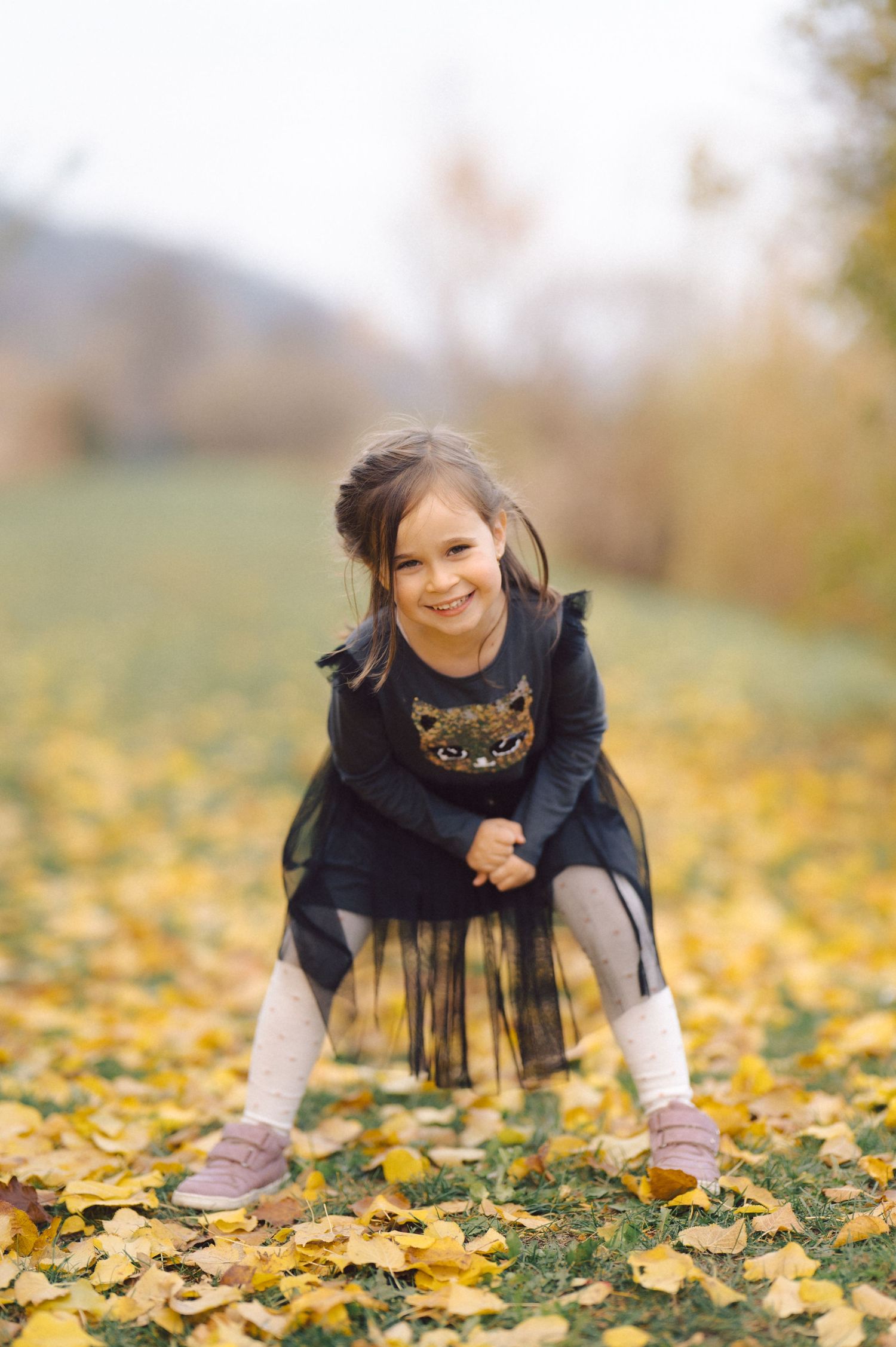 Child in black dress crouches among fallen yellow autumn leaves with joyful expression.