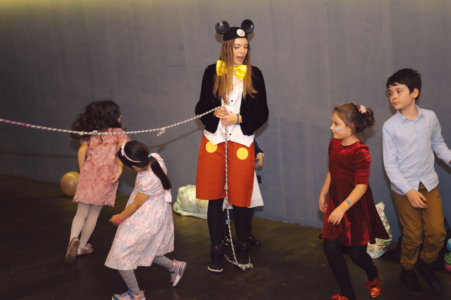 Children play tug-of-war with someone dressed as Mickey Mouse at a party.