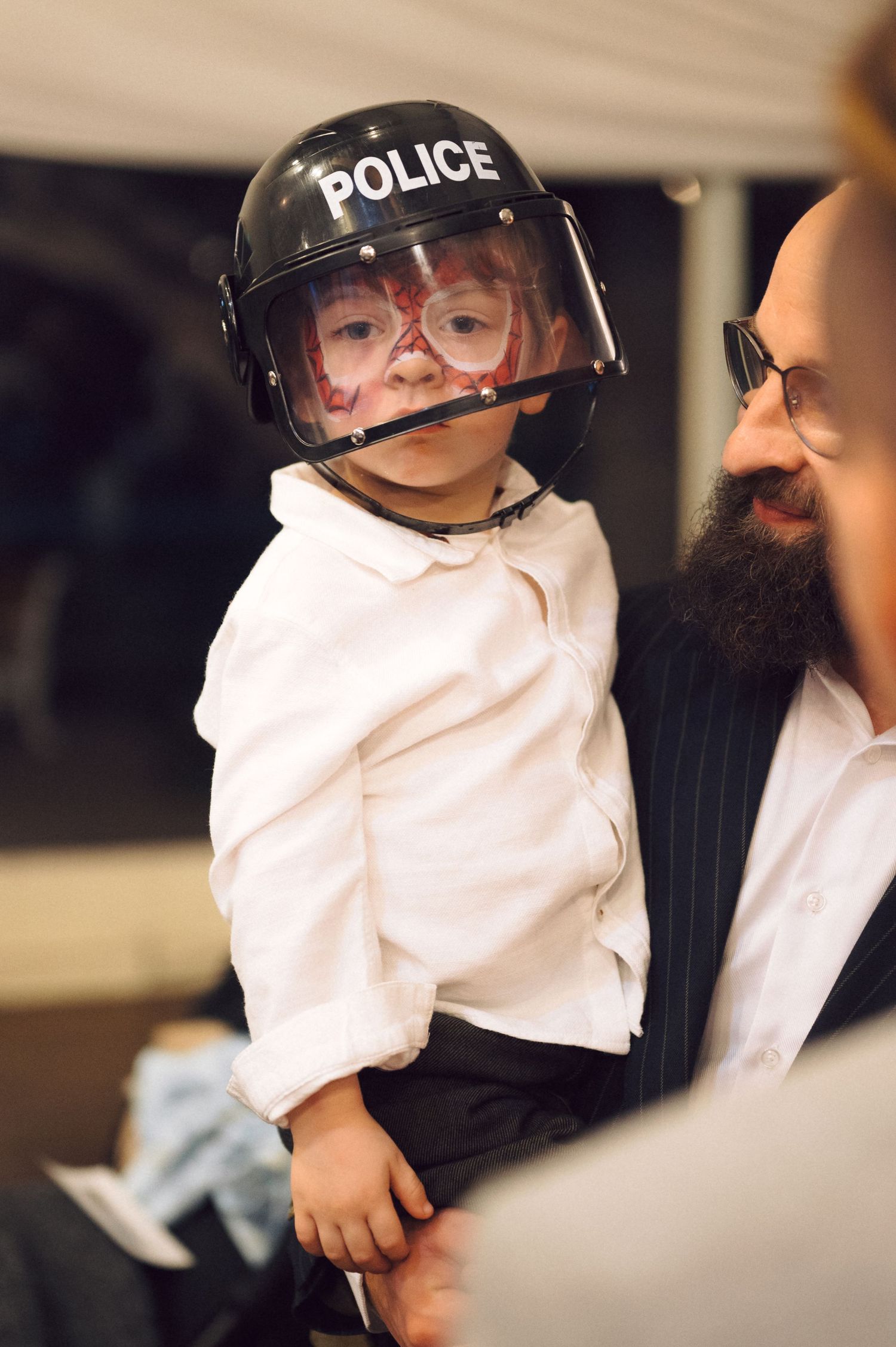 Small child wearing a police helmet and glasses stands next to someone with a beard.