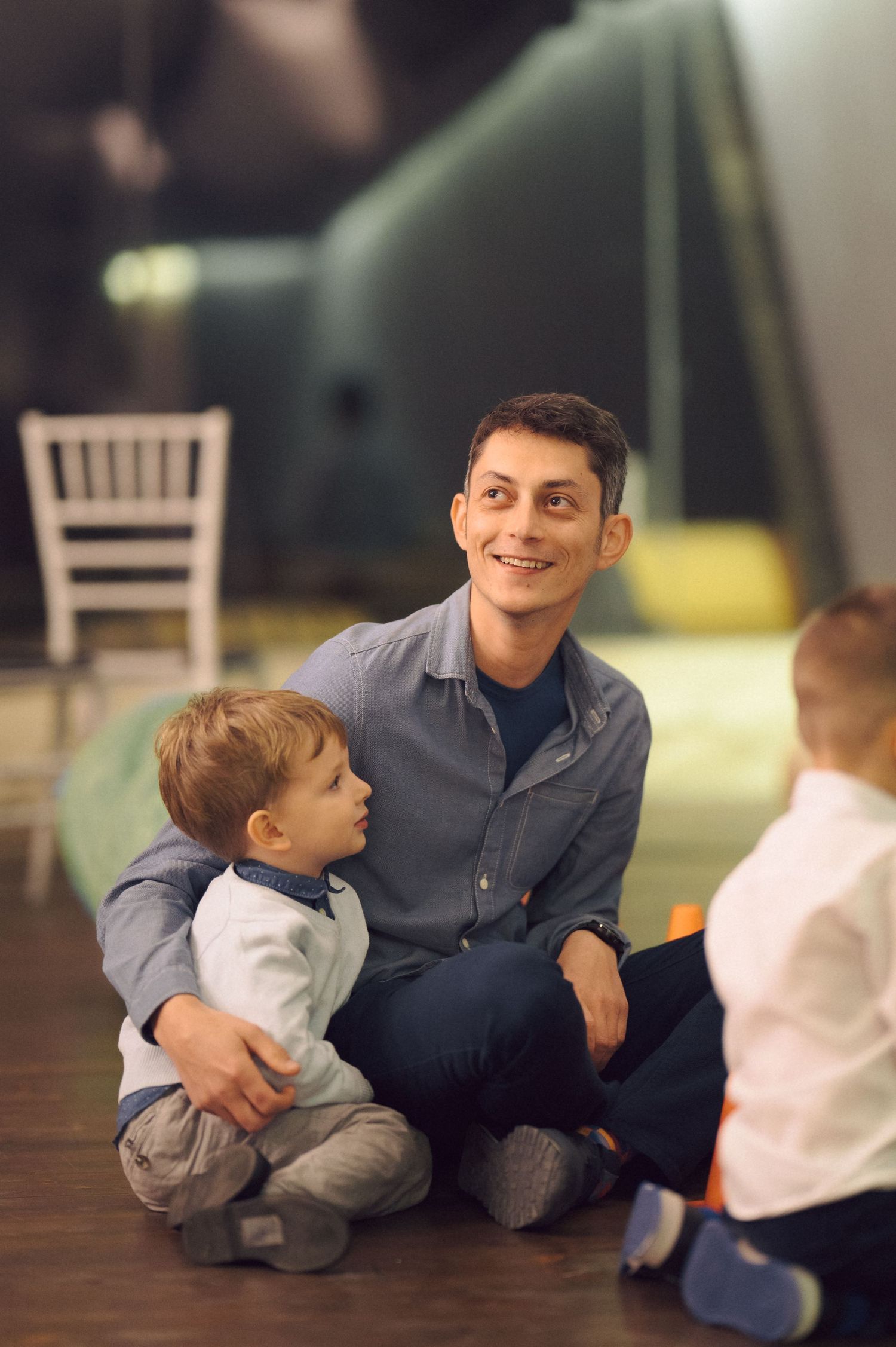Father and young children sitting together on wooden floor at indoor event.