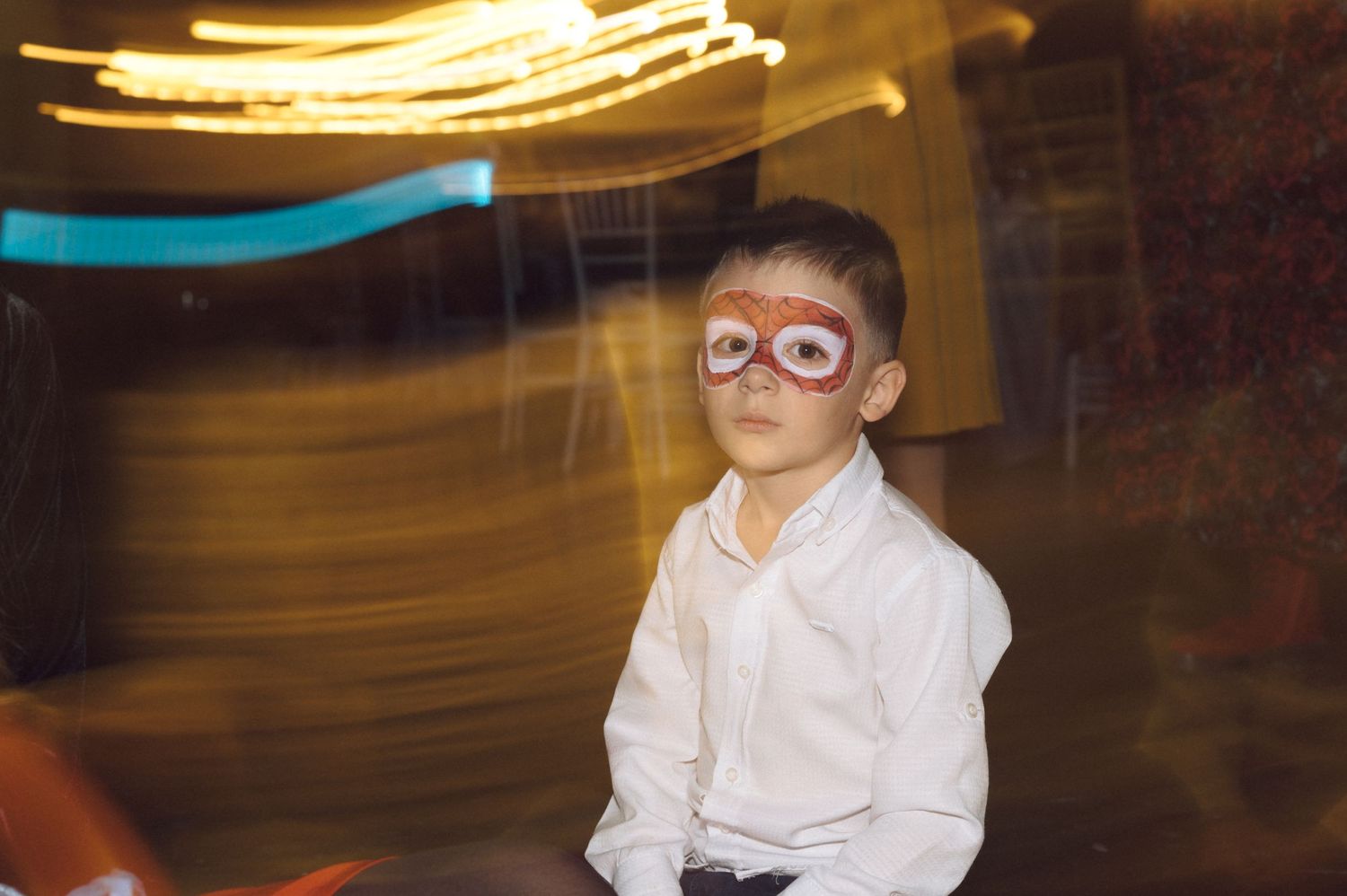 Young child in white shirt and glasses smiles at indoor party with blurred lights.
