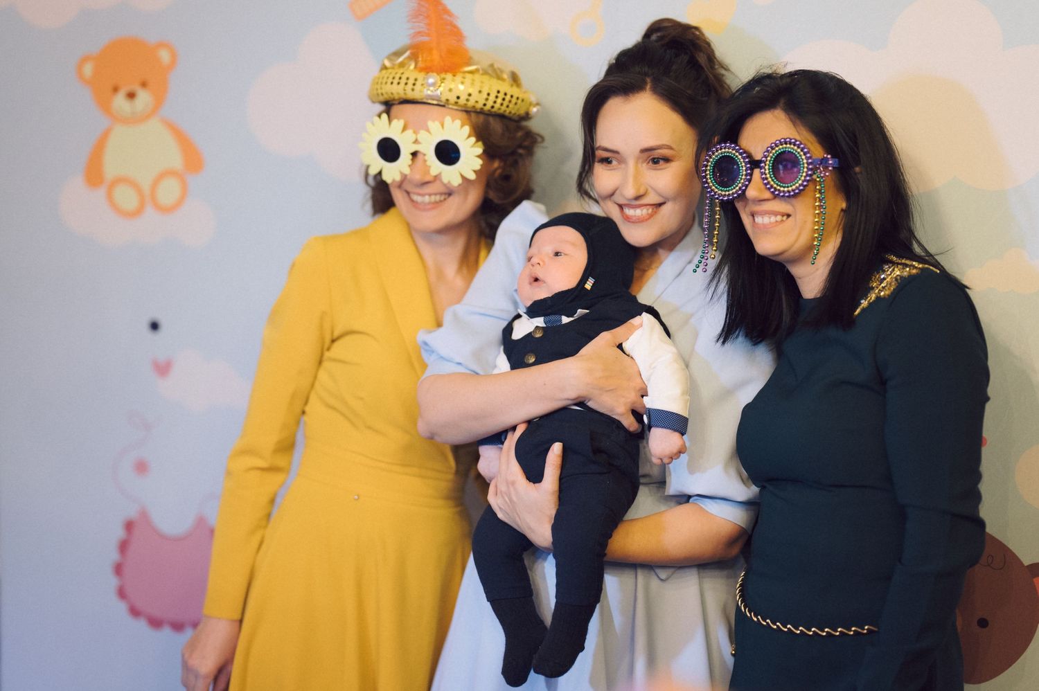 Three guests and a baby pose together at a celebration wearing festive outfits and novelty sunglasses.
