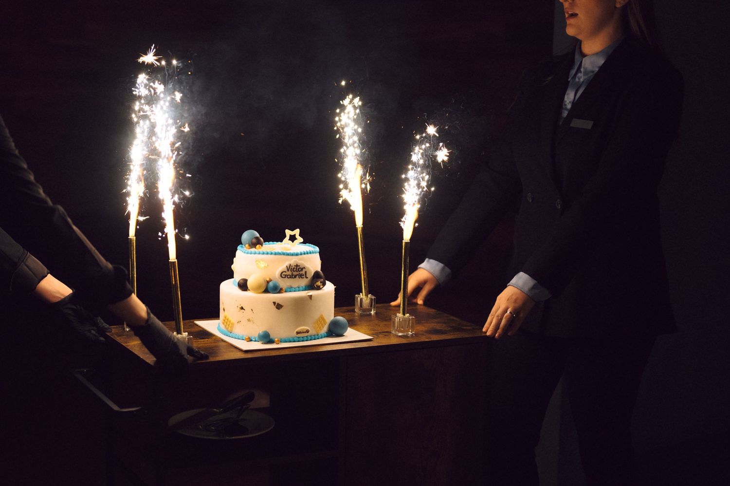 A birthday cake with sparkler fountains is presented in a dimly lit room.