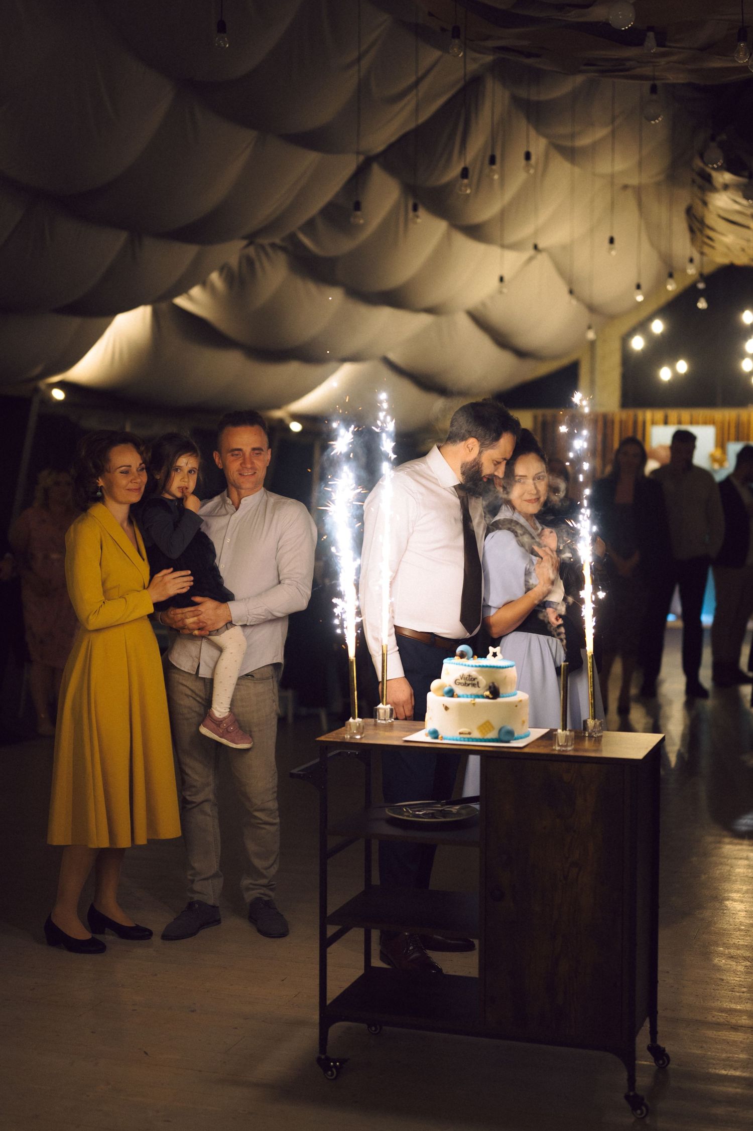 Guests gather around a cake with sparklers under draped fabric ceiling decor.