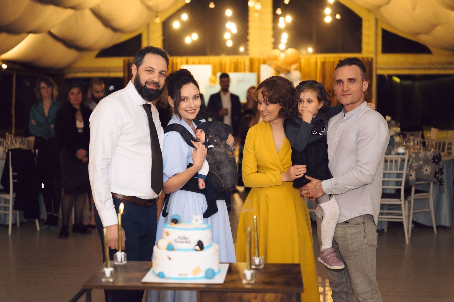 A group poses with a blue and white tiered celebration cake at an indoor event.