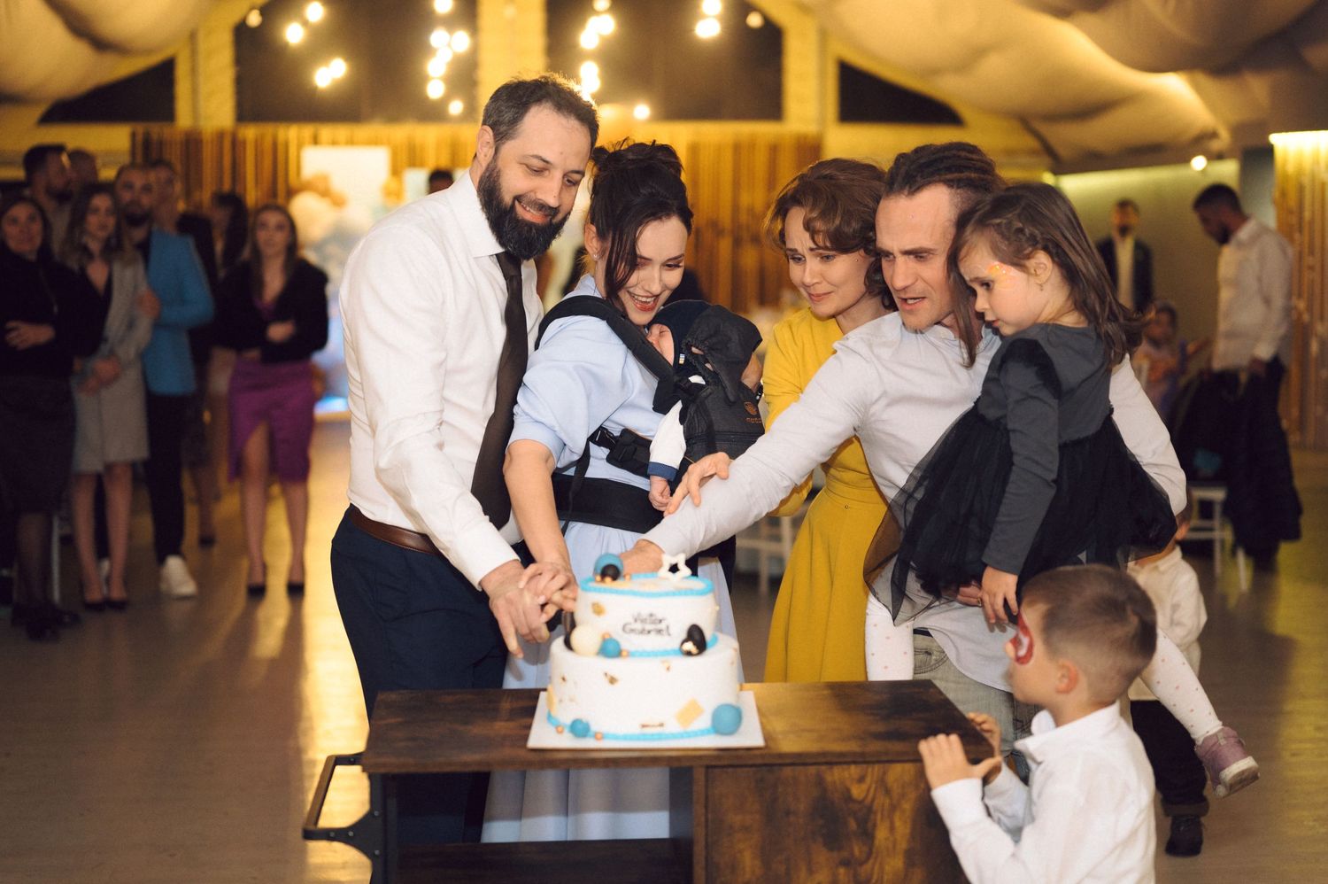 Multiple guests gather around to cut into a decorated tiered cake at a celebration.