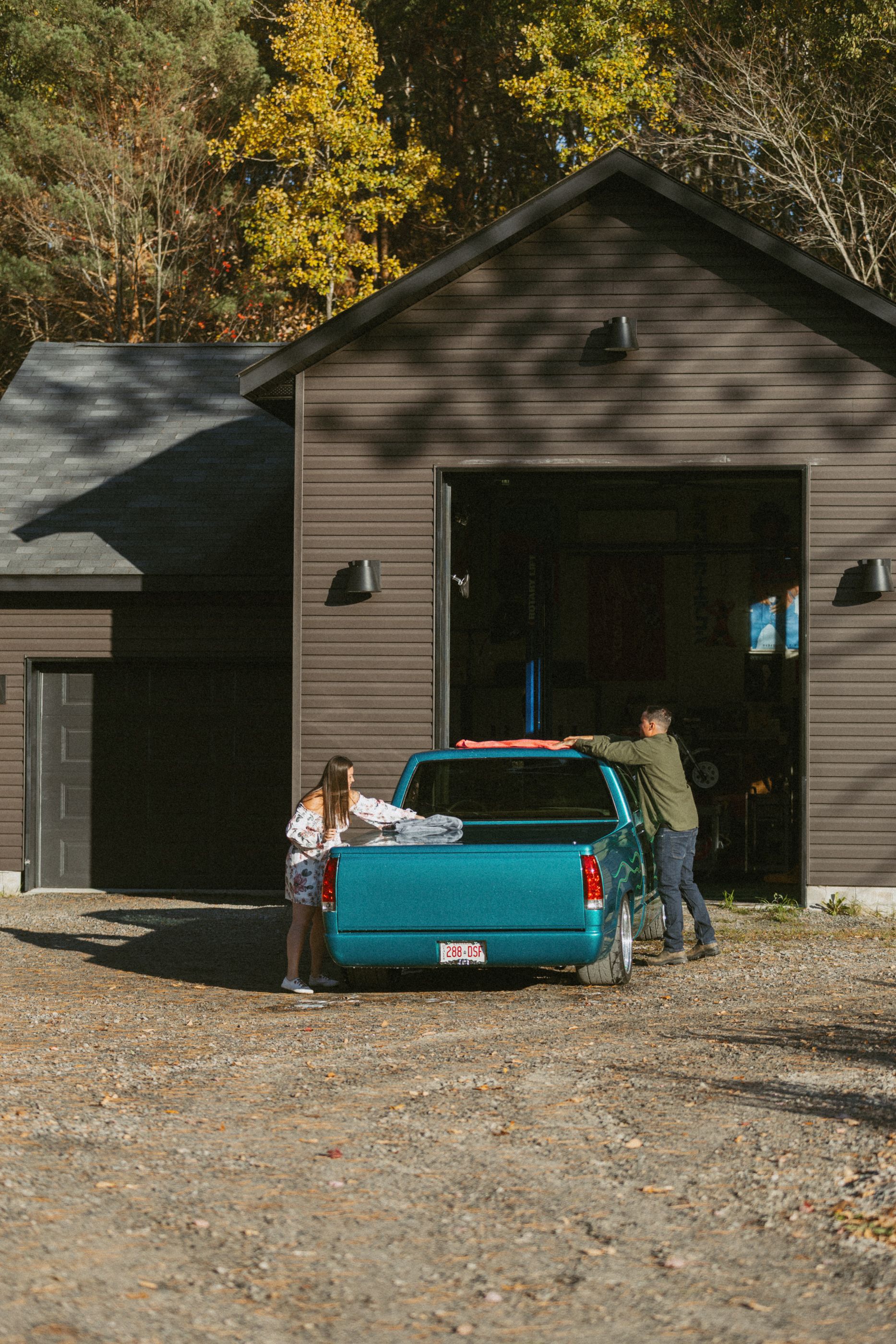 A couple stands beside a turquoise pickup truck in front of a brown garage on an autumn day.