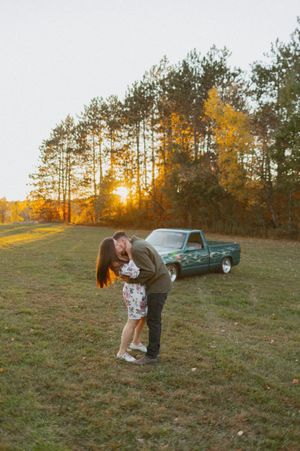 A romantic sunset engagement photo with a vintage pickup truck parked in a field surrounded by autumn trees.
