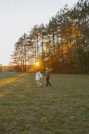 A couple walks hand in hand across a field at sunset with tall pine trees in the background.